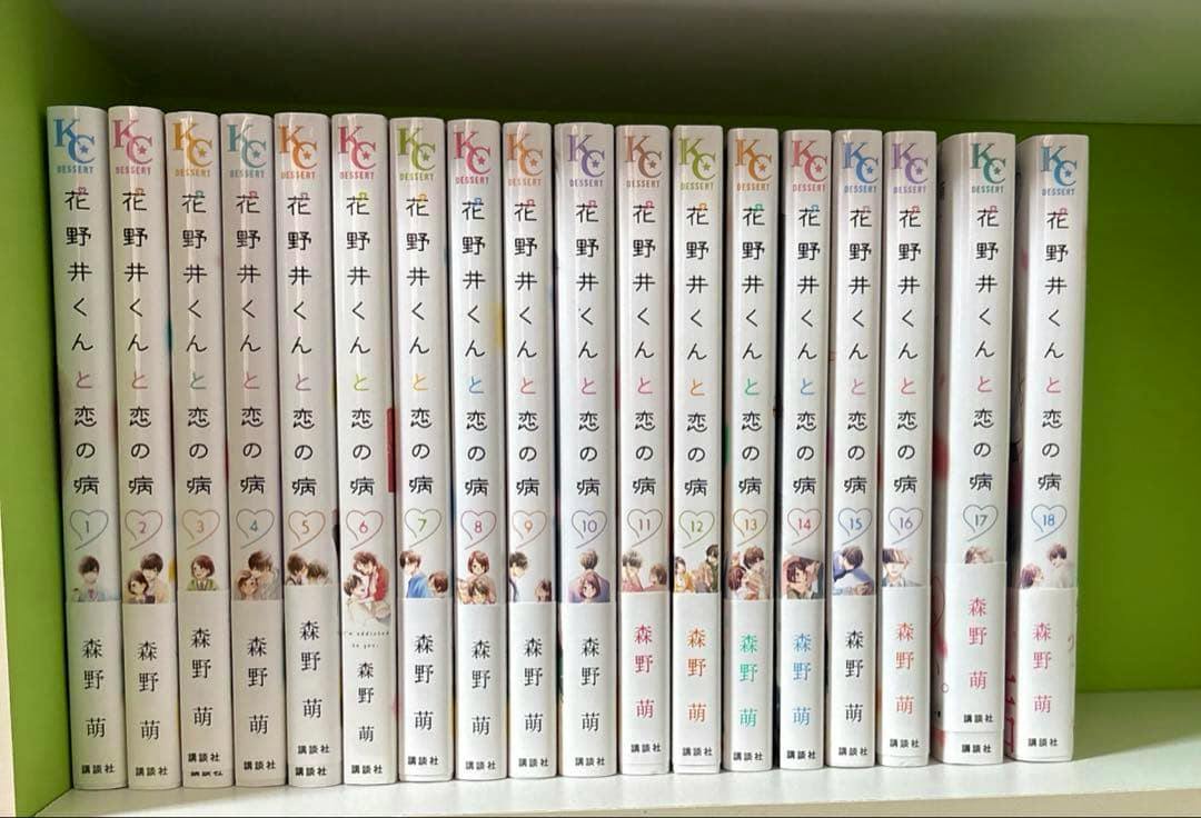 Eighteen volumes of the manga '花野井くんと恋の病' (volumes 1–18) lined up with spines visible on a green shelf.