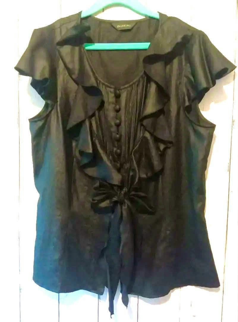 Front view of a black short-sleeve women's blouse with ruffles, fabric-covered buttons, and a tied front bow hanging on a hanger.