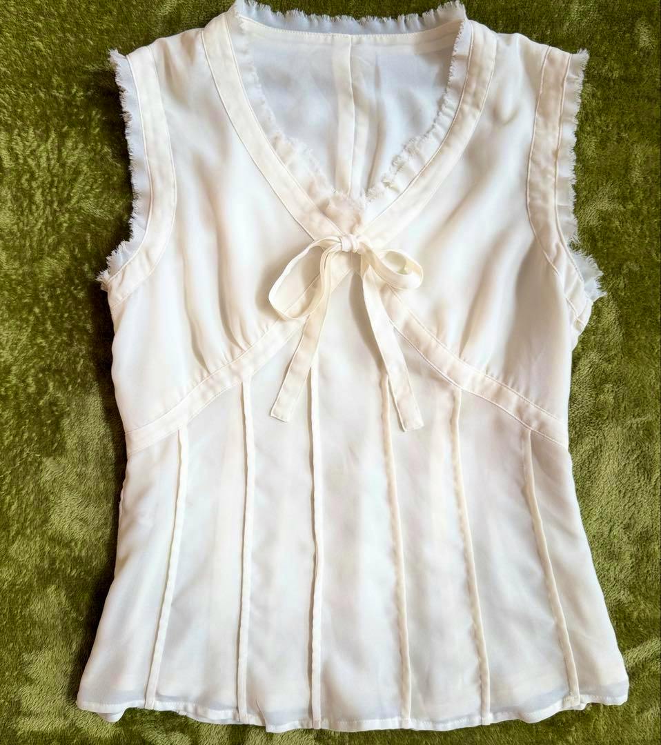 Ivory INGEBORG sleeveless women's polyester blouse (size M) with V-neck, ribbon tie, and frill trim laid flat on a green background.