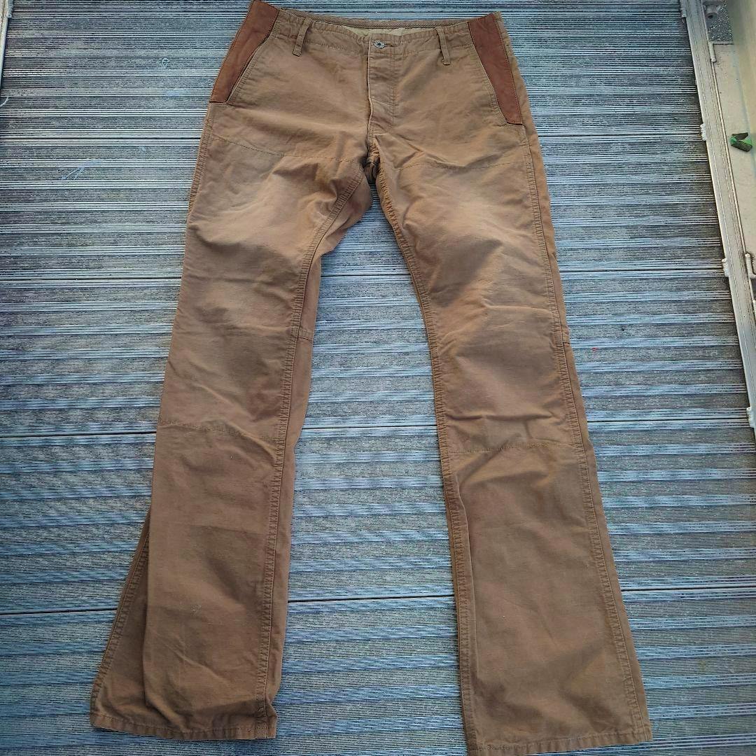 Front view of brown ISAMU KATAYAMA BACKLASH chino pants laid flat on a textured surface.