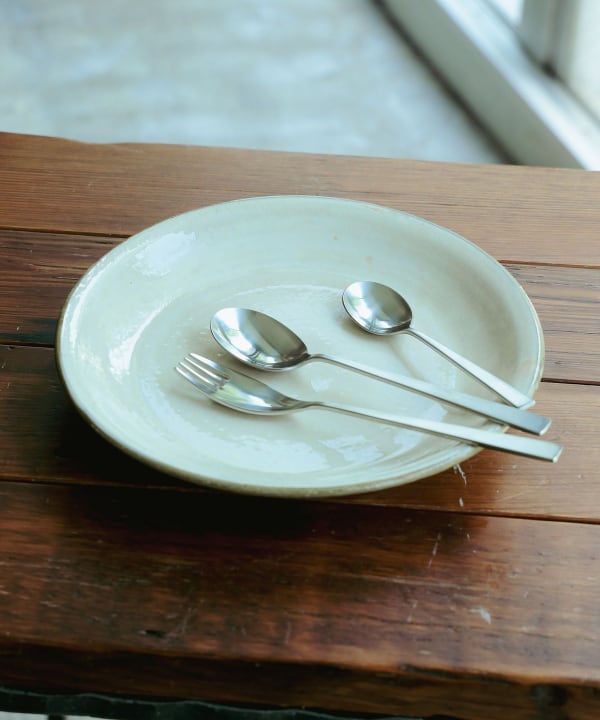 A metal fork and two spoons arranged on a ceramic plate on a wooden surface.