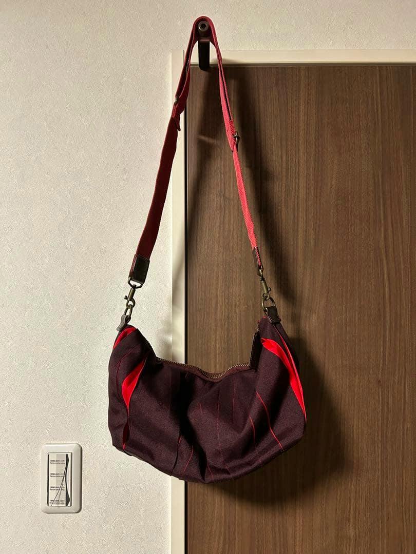 Dark purple and red Issey Miyake shoulder bag hanging on a door hook, showing the full bag and adjustable red strap.
