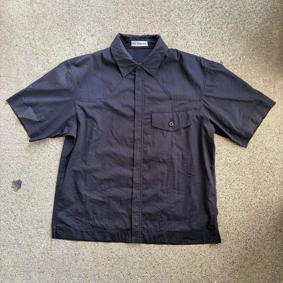 Black men's short-sleeve zip-up shirt laid flat on a textured surface, showing collar and chest pocket.