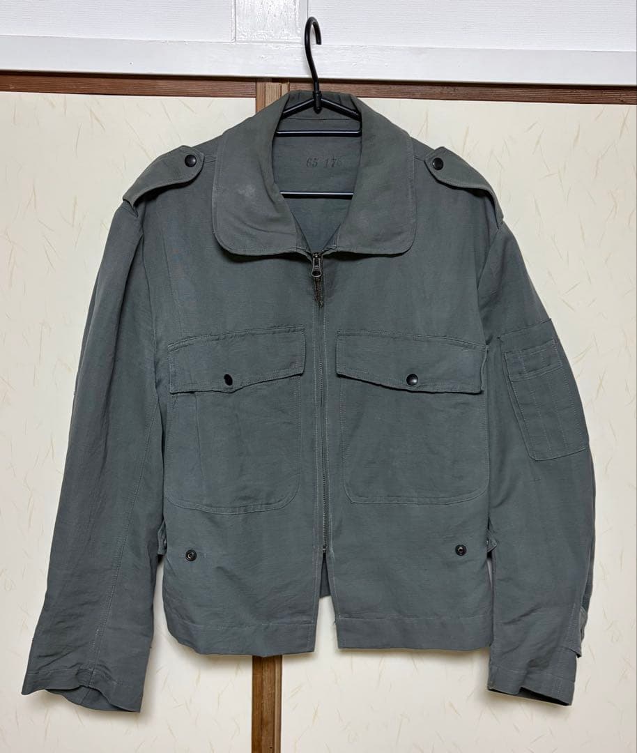 Front view of the Italian military 1960s flight jacket (mens cotton woven) hanging on a hanger, showing a center zipper, two flap chest pockets, epaulets, and buttoned hem tabs.
