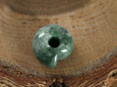 Green-and-white jadeite magatama pendant with a drilled hole resting on a wooden surface.