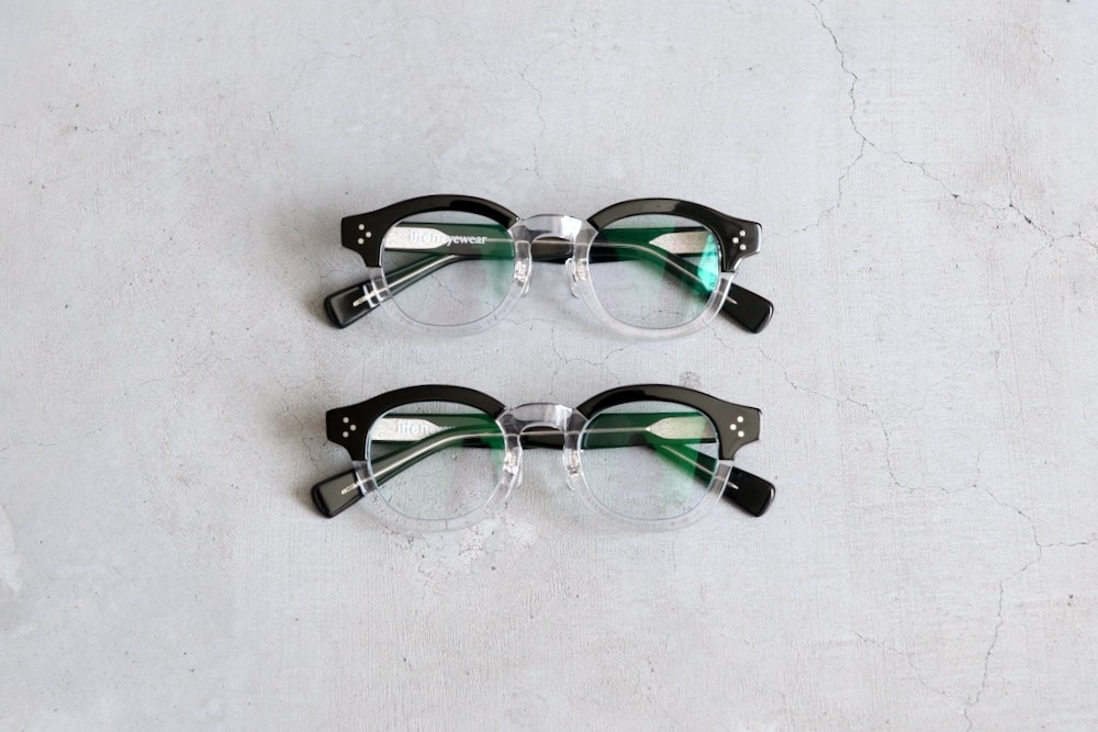 Two itten. "eye wear 002" corrective eyeglasses with dark upper rims and clear lower rims laid flat on a light background.