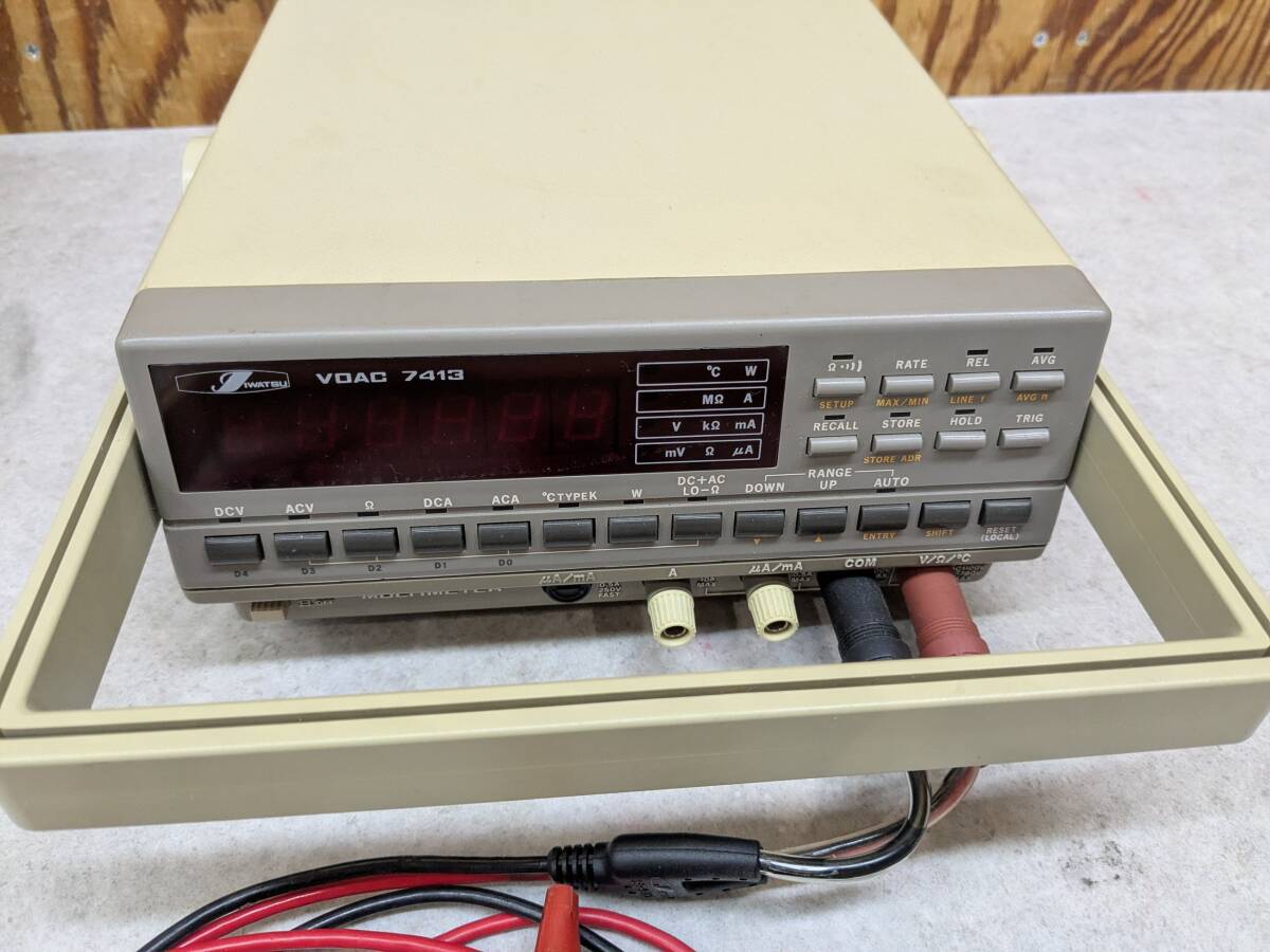 Front view of the IWATSU VOAC 7413 digital multimeter showing the display, front panel buttons, and input terminals with attached test leads.