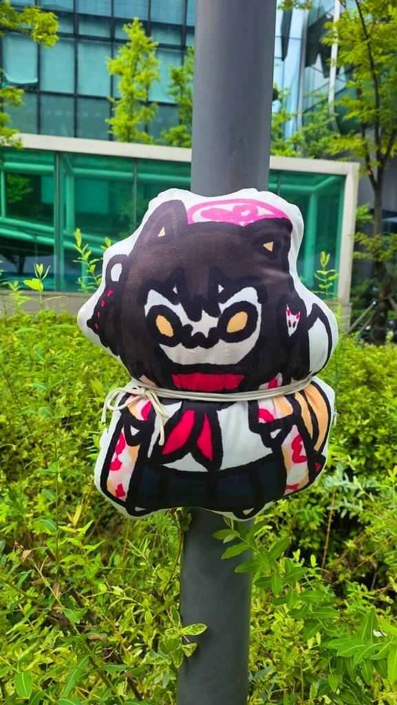 A 35cm Izuna plush toy with dark hair and red-accented outfit tied to a pole outdoors.