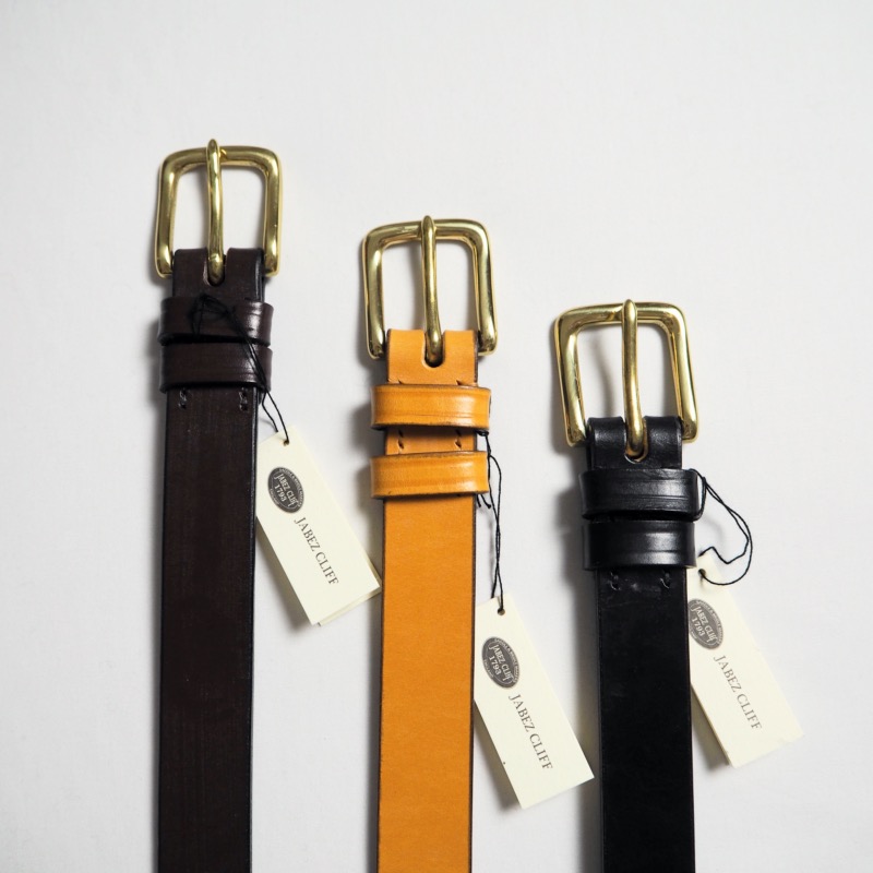 Three cow leather belts in chestnut, tan, and black with metal buckles and hang tags laid on a white background.