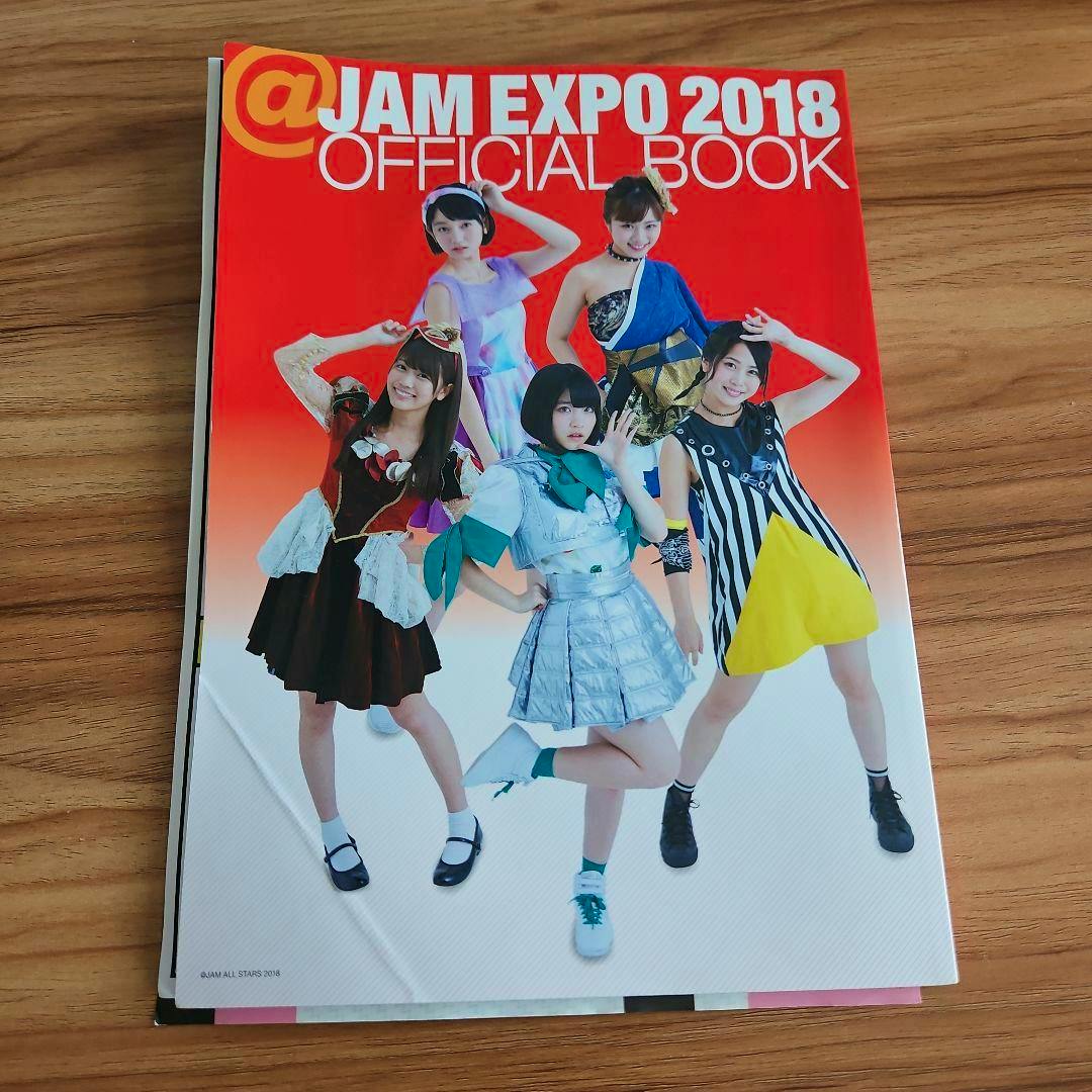 Front cover of the @JAM EXPO 2018 official book showing performers in colorful costumes on a wooden surface.