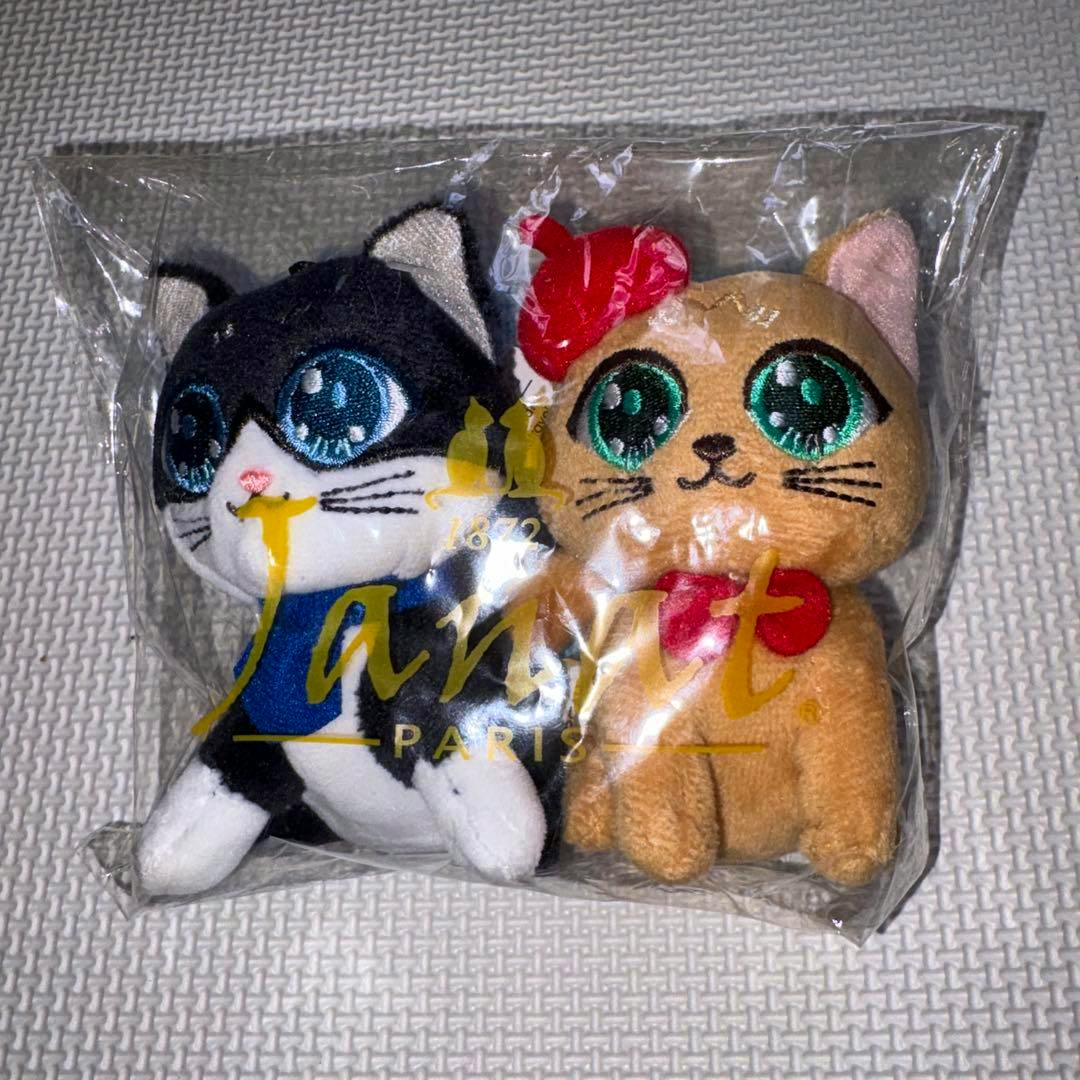 Two Janat plush cat mascot toys visible in clear plastic packaging, one black-and-white and one tan with a red bow.