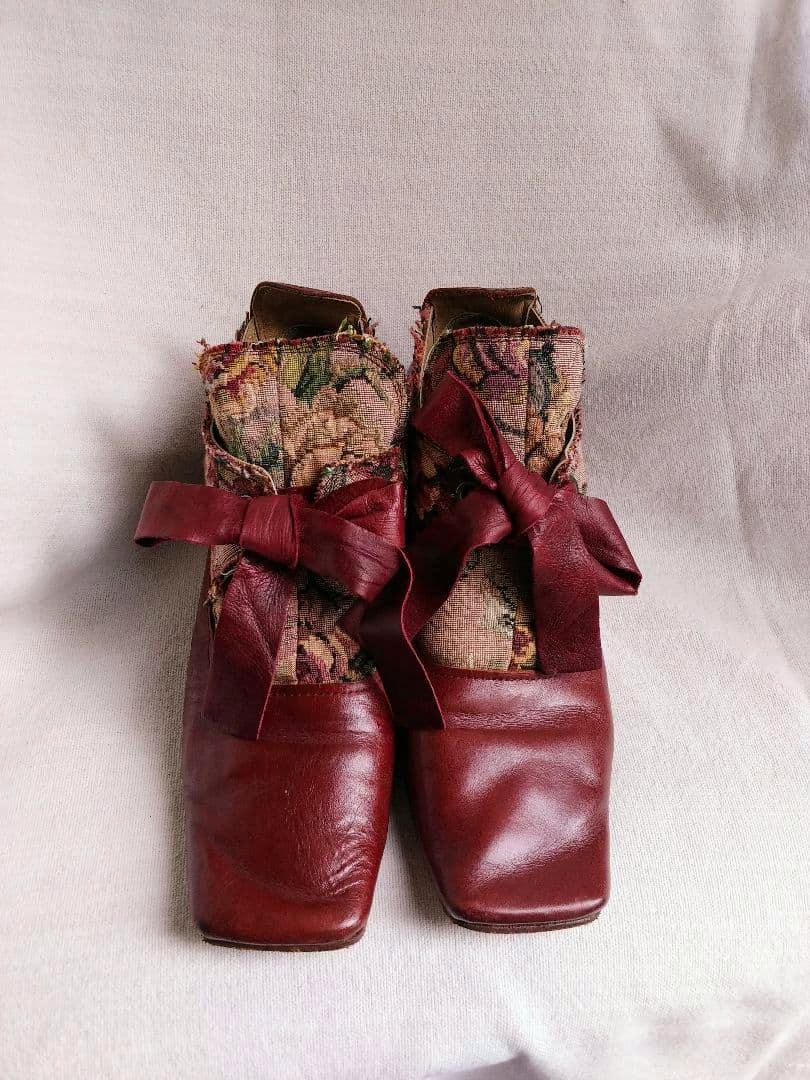 Front view of a pair of bordeaux leather loafers with gobelin tapestry panels and leather bows on a pale fabric background.
