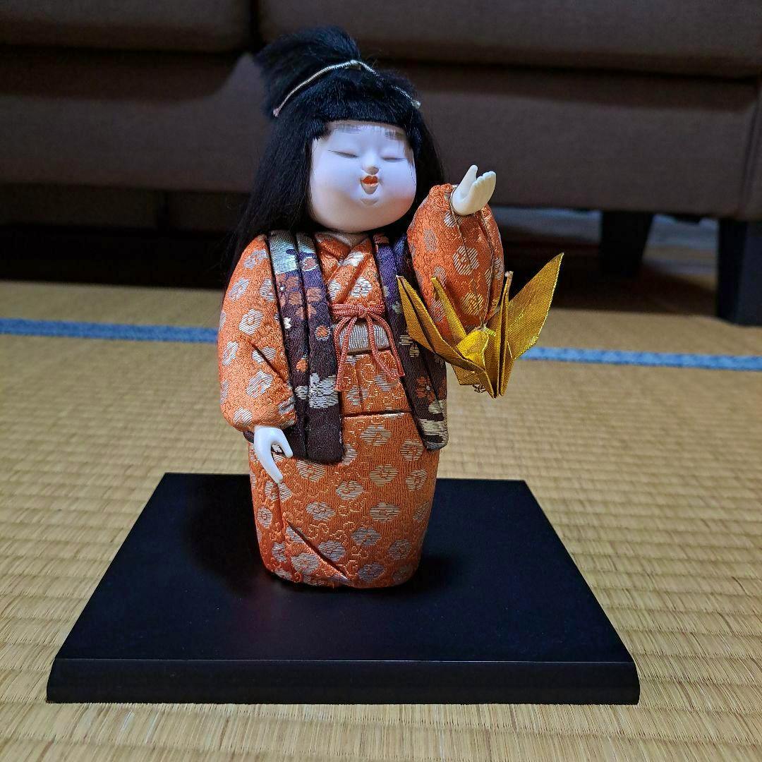 Front view of the Japanese doll in an orange kimono holding a golden origami crane on a black display base.