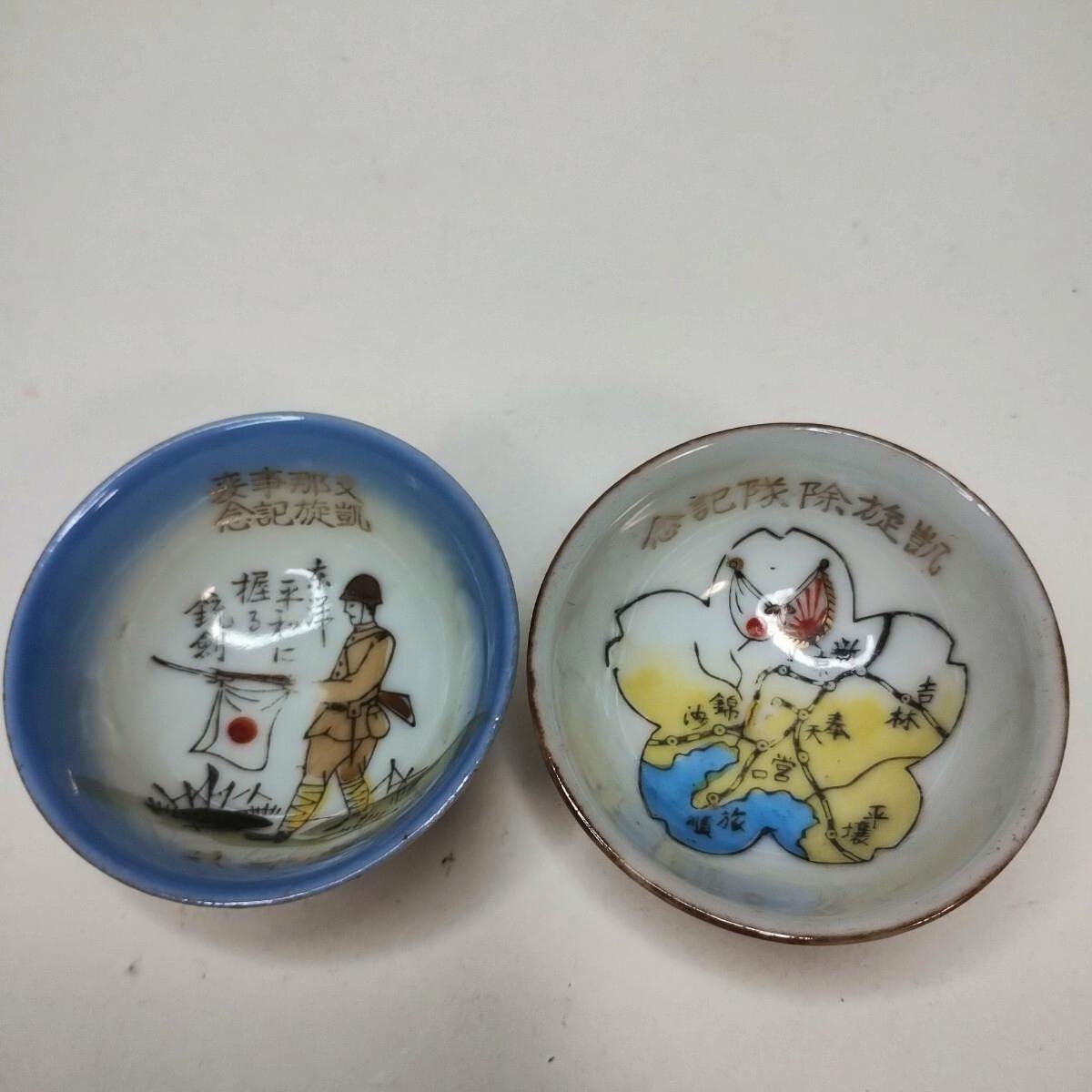 Two small ceramic commemorative military cups showing a soldier and a map design, photographed from above on a white surface.