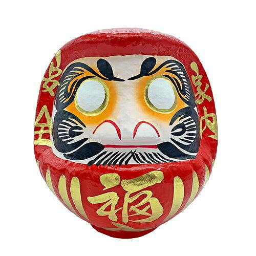 Front view of a red Japanese daruma figure with gold and black facial decorations on a white background.
