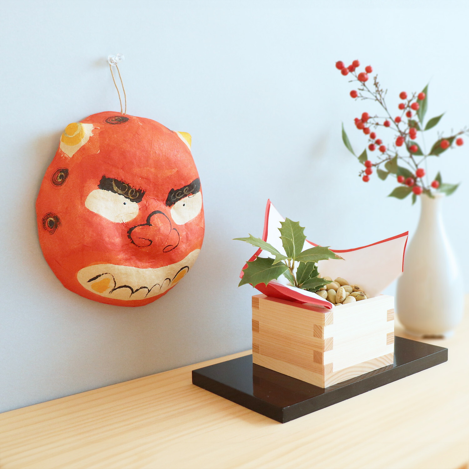 Red papier-mâché oni (red demon) face ornament hanging on a wall next to a wooden box of beans and a vase.