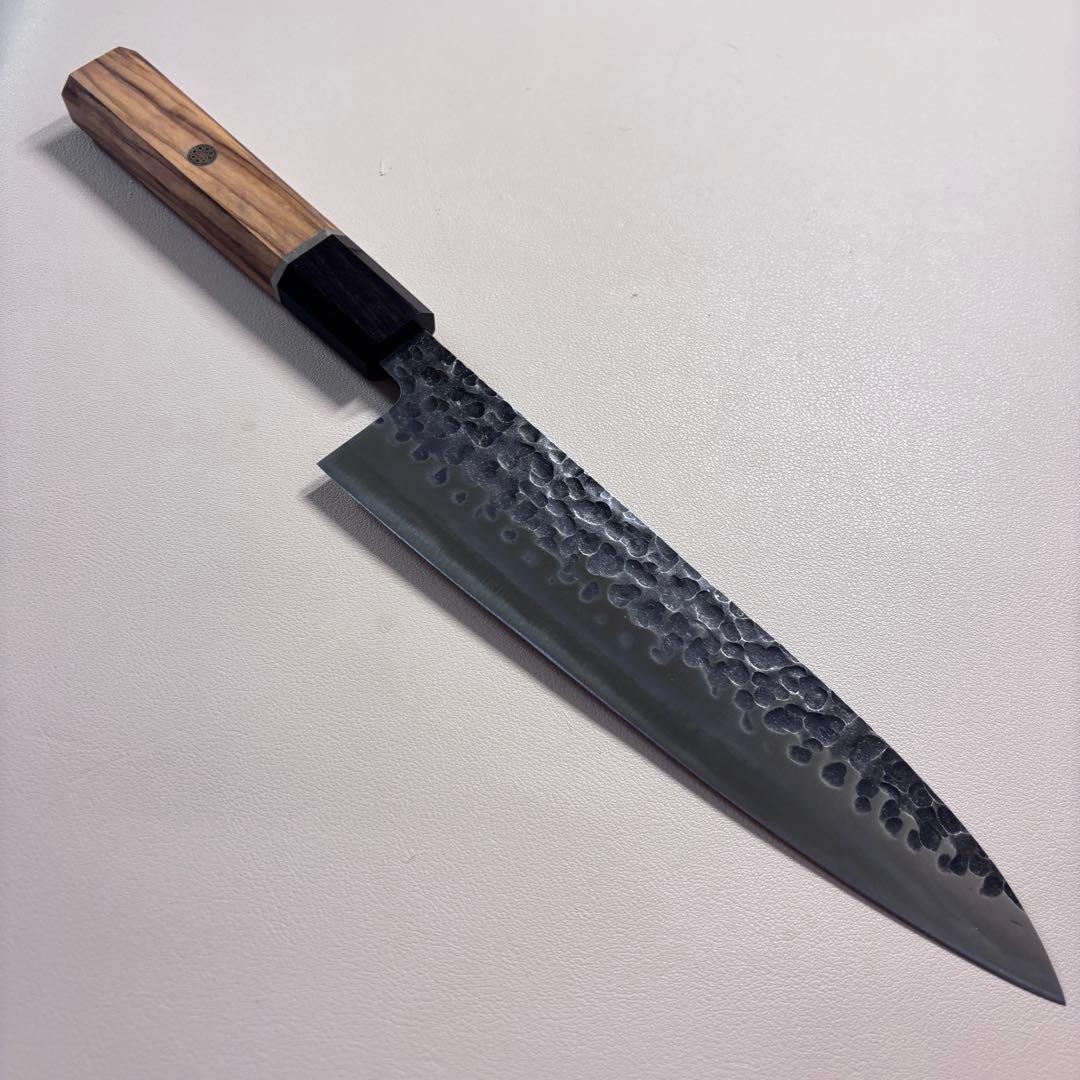 A 210mm 440C stainless chef knife with a hammered blade texture and an octagonal wooden handle lying on a light surface.