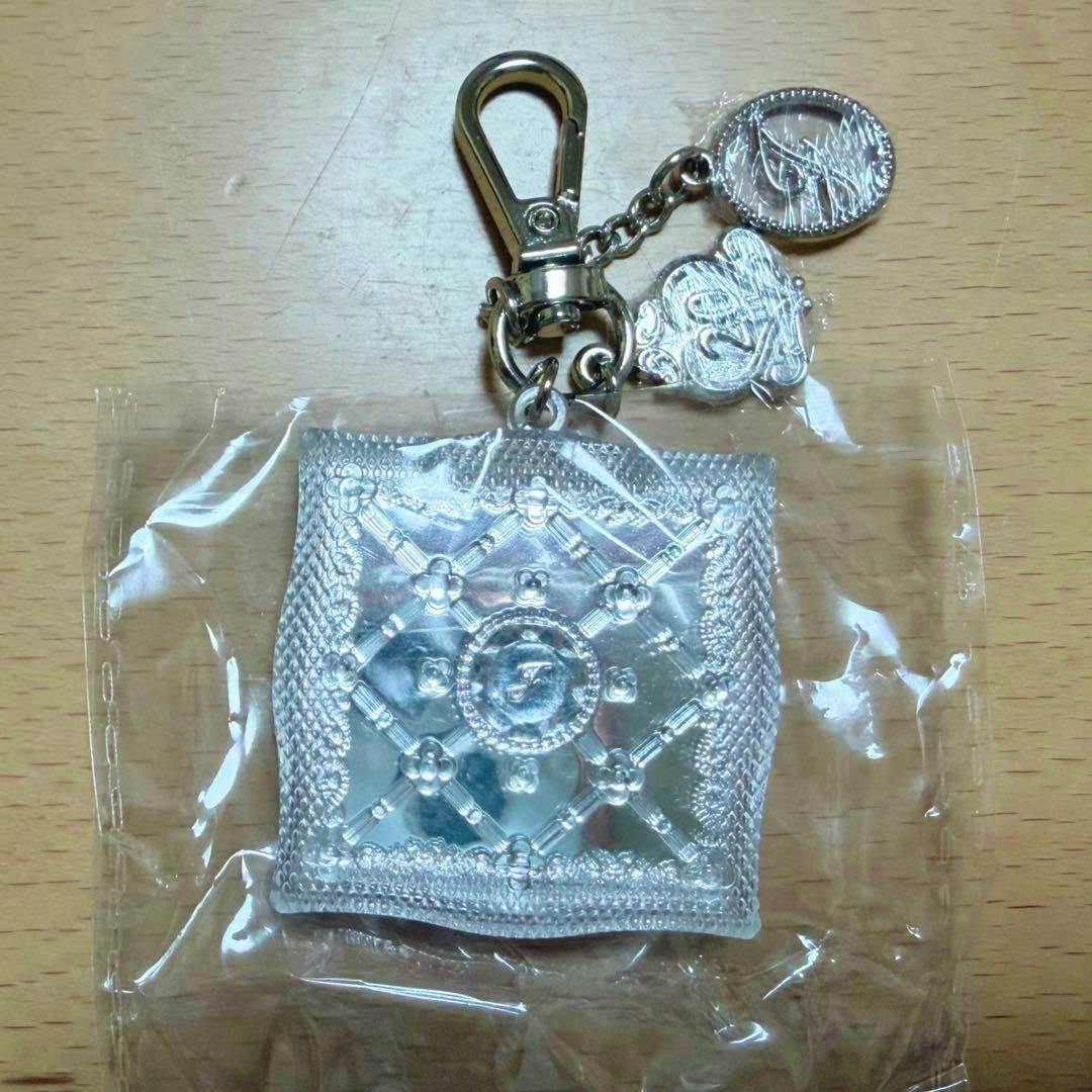 Silver-toned mini compact bag charm with a clasp and decorative charms, shown in clear plastic packaging on a wooden surface.