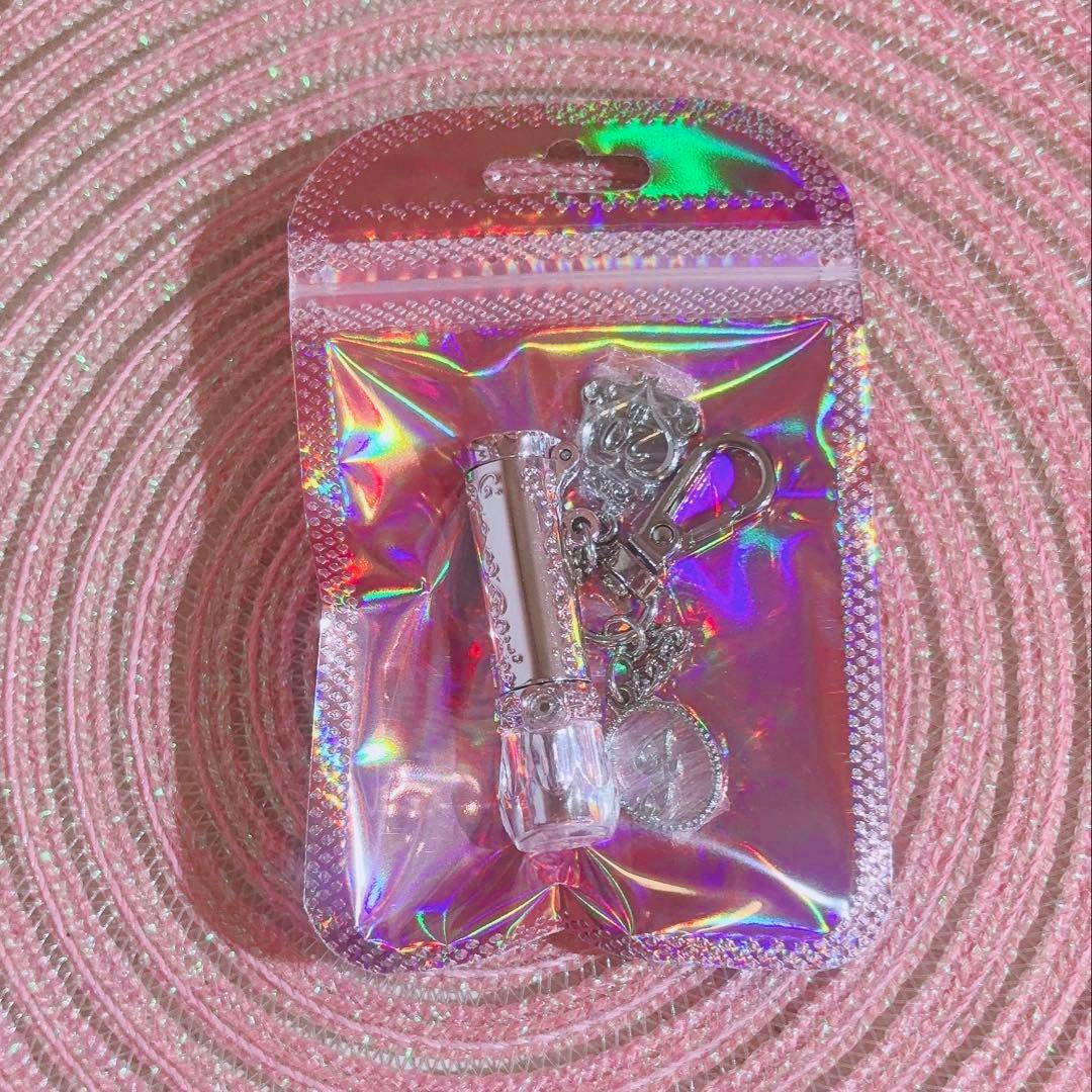 Packaged keychain shaped like a lip balm tube inside a holographic pouch on a pink textured surface.
