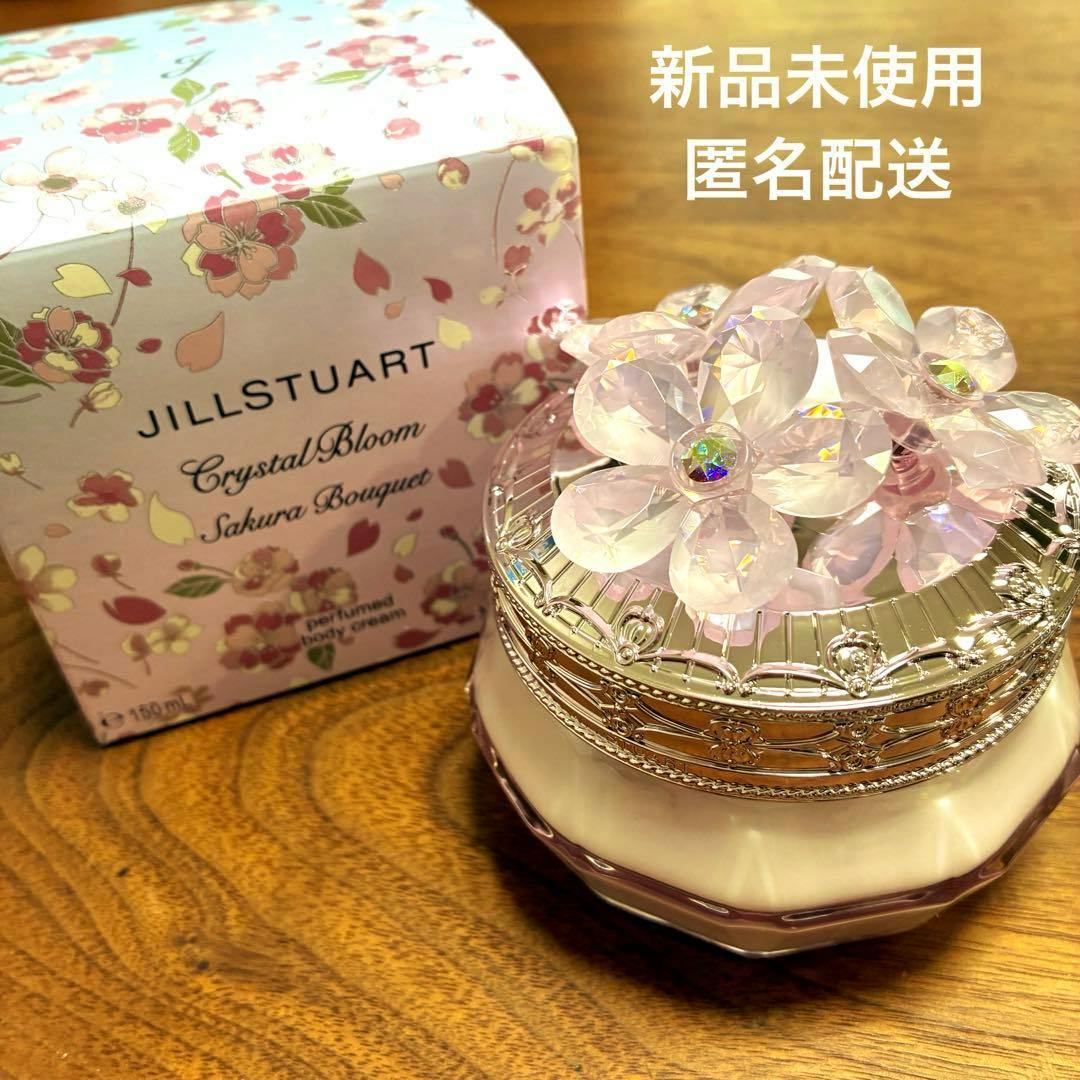 A JILLSTUART Crystal Bloom Sakura Bouquet perfumed body cream jar with a decorative flower-topped lid beside its floral box on a wooden surface.