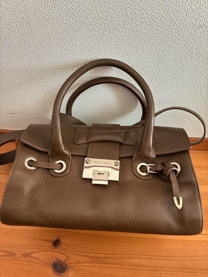 Brown Jimmy Choo leather handbag with two rounded top handles, a front metal lock plate, and grommet eyelets with a leather strap, shown on a wooden surface.