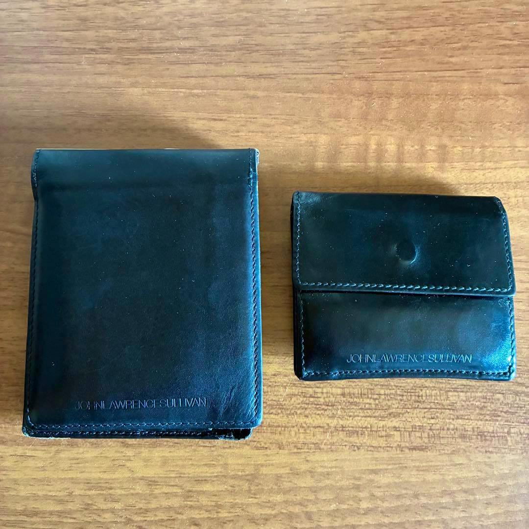 A black JOHN LAWRENCE SULLIVAN bi-fold wallet placed on a wooden surface next to a smaller black leather accessory.