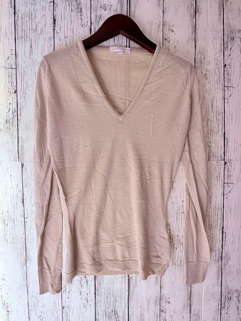 Beige John Smedley V‑neck knit sweater hung on a wooden hanger against a white wood background.