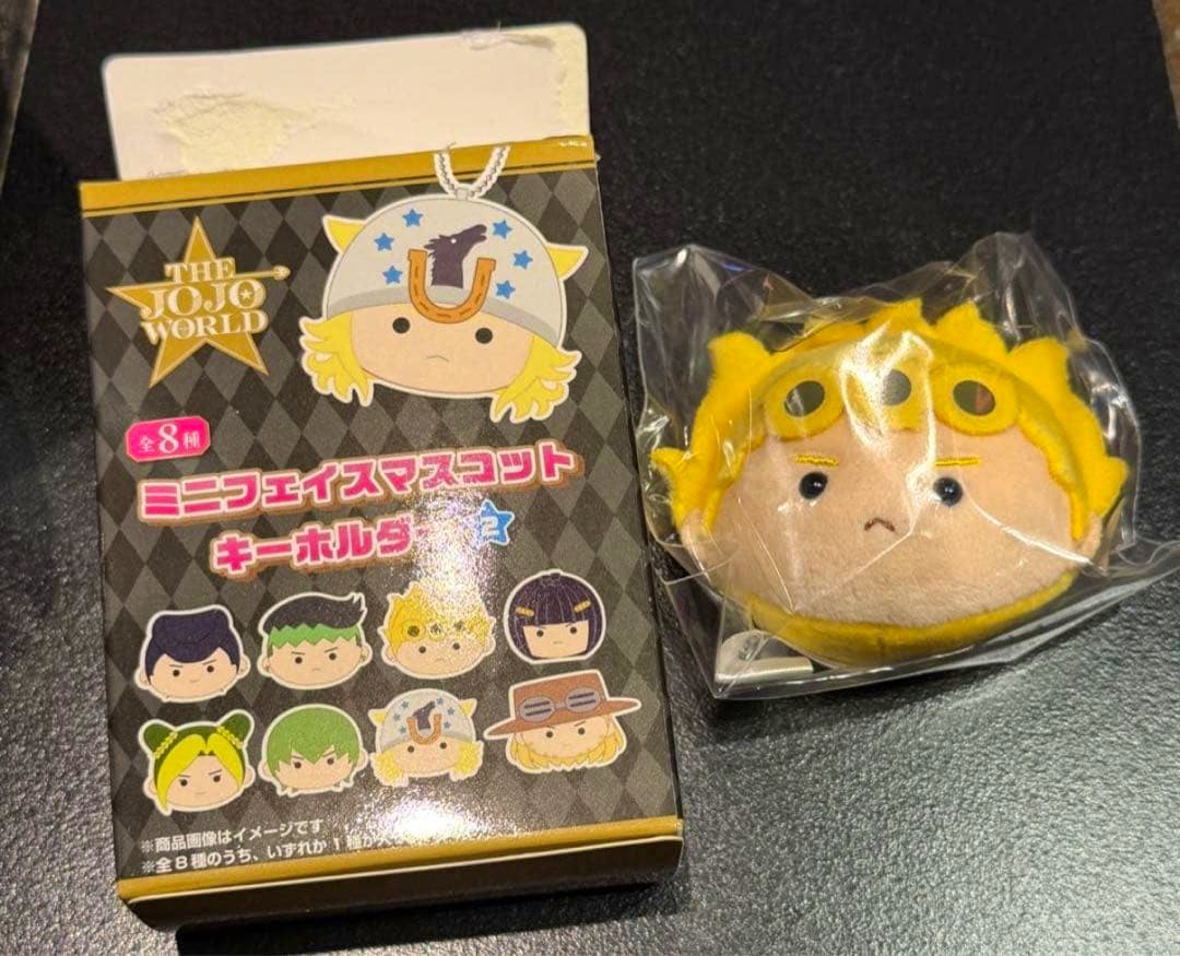 A Giorno mini face plush mascot in clear plastic packaging beside its JoJo World product box showing other character designs.
