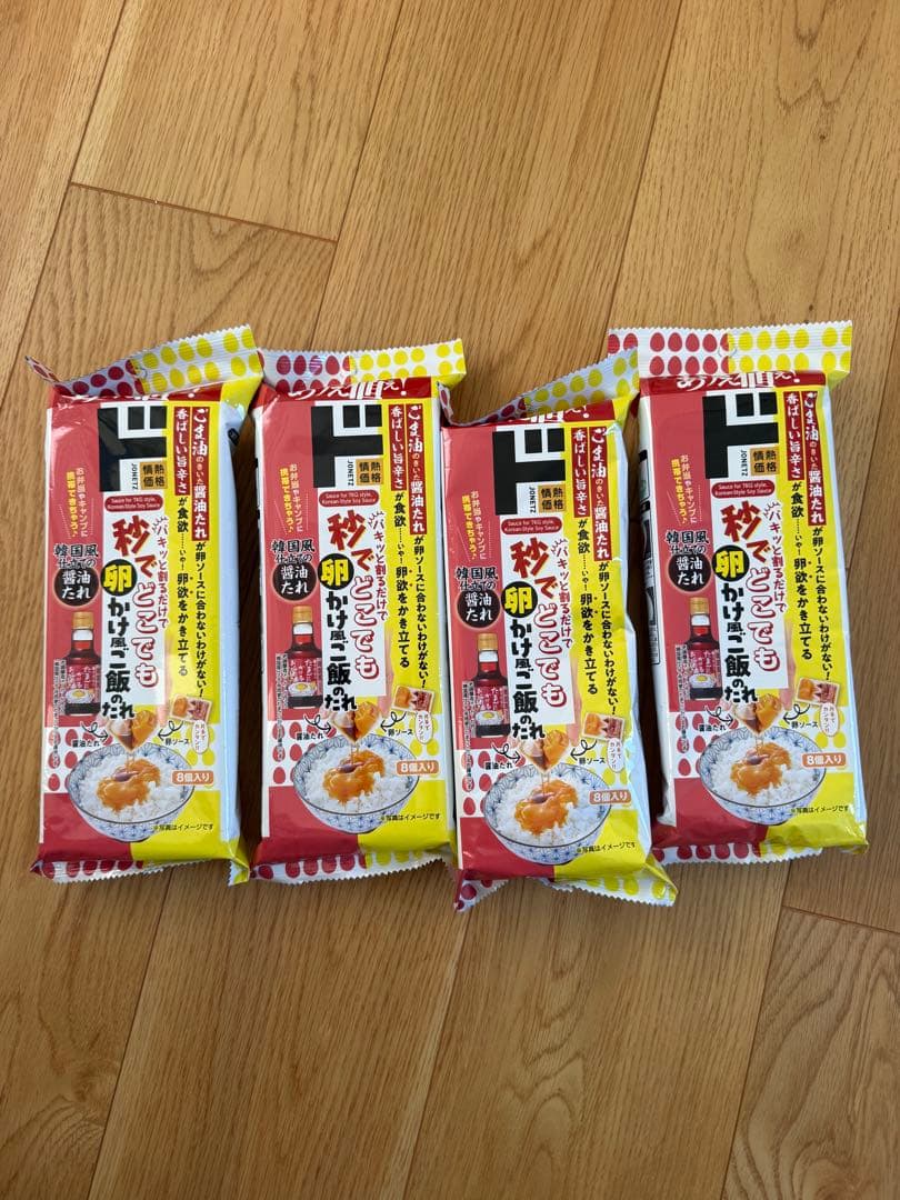 Four packaged units of the rice seasoning “情熱価格 秒でどこでも卵かけ風ご飯のたれ” arranged on a wooden floor with front labels visible.