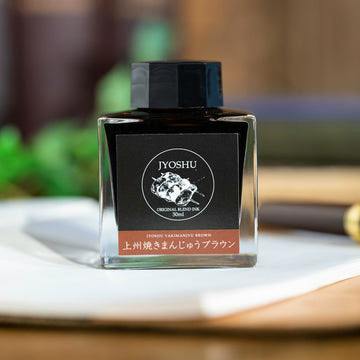 Close front view of the fountain pen ink bottle (上州焼きまんじゅうブラウン) placed on a notebook.