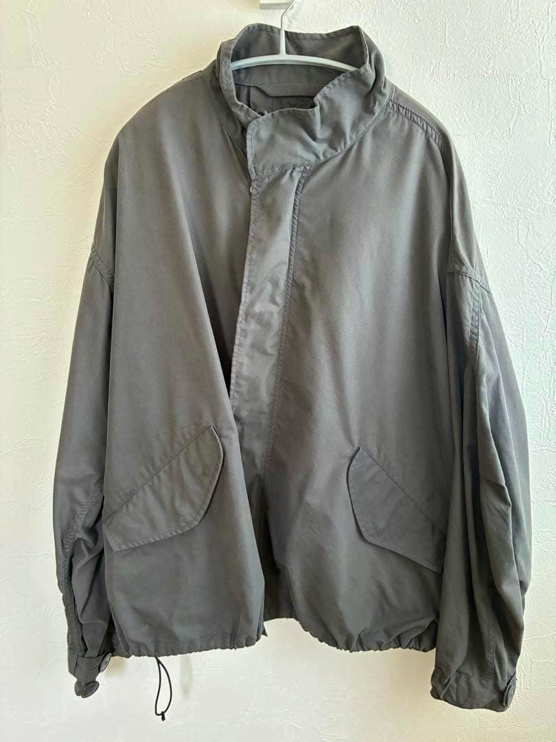 Front view of a gray men's nylon jacket hanging on a hanger, showing flap pockets and a drawstring hem.