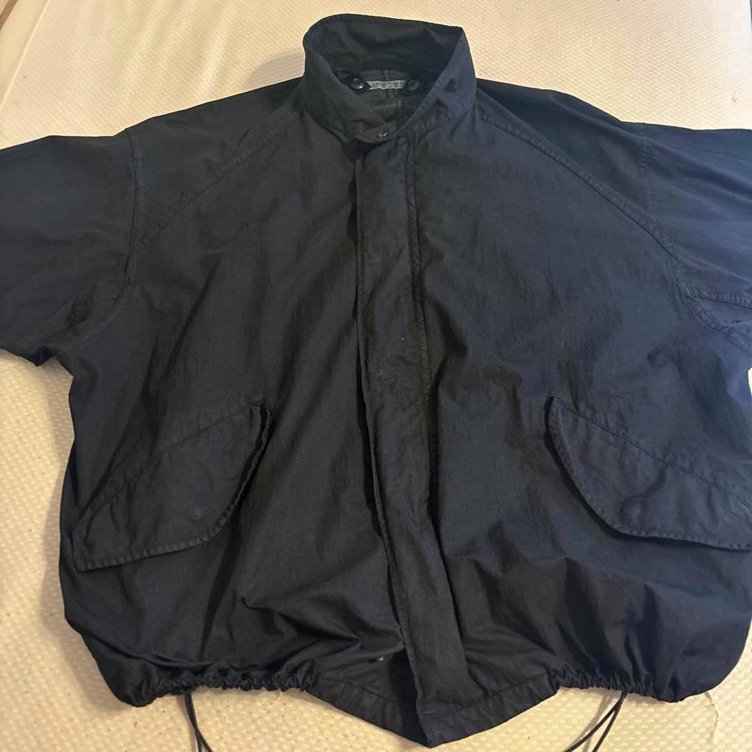 Dark nylon men's jacket laid flat showing the front, flap pockets, high collar, and drawstring hem.