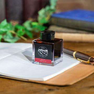 A bottle of fountain pen ink (KOKIA Red) sitting on an open notebook with a fountain pen nearby.