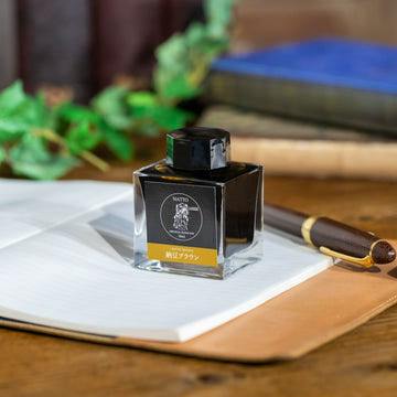 Square bottle of fountain pen ink labeled "納豆ブラウン" on an open notebook with a fountain pen and books in the background.