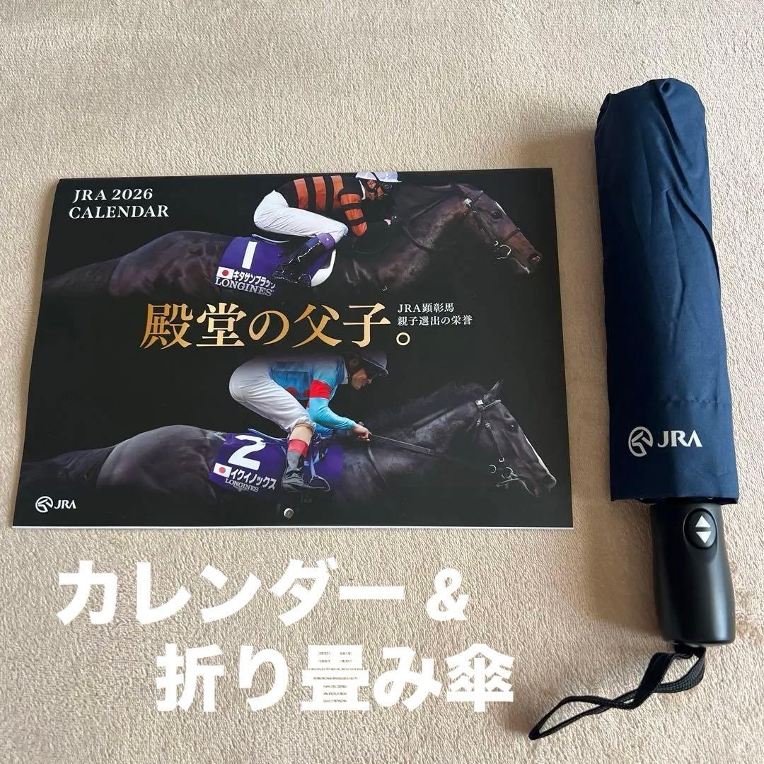 A folded navy JRA-branded folding umbrella beside a JRA 2026 calendar on a beige surface.
