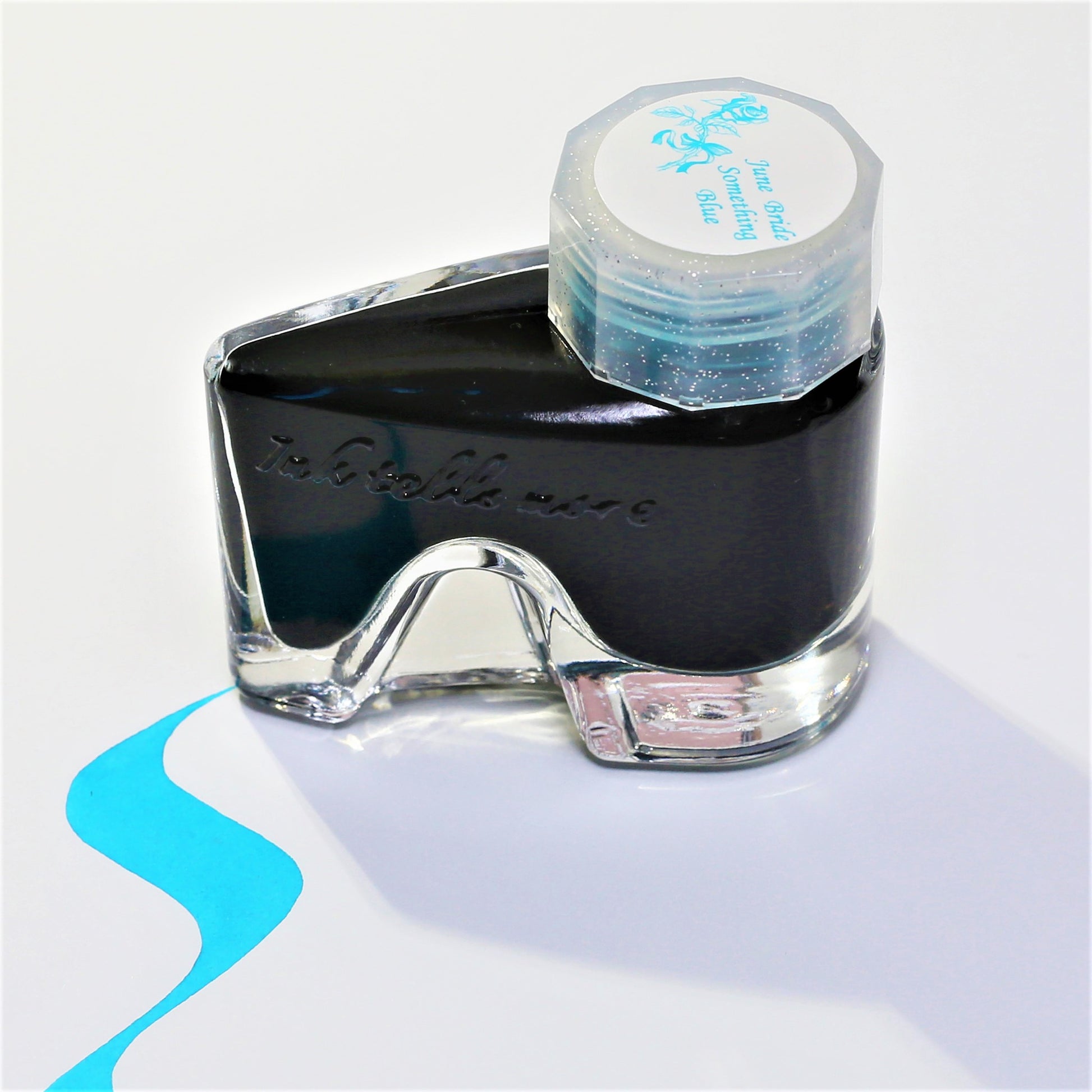 A glass fountain pen ink bottle of June Bride Something Blue Ink with a blue ink swatch on a white background.