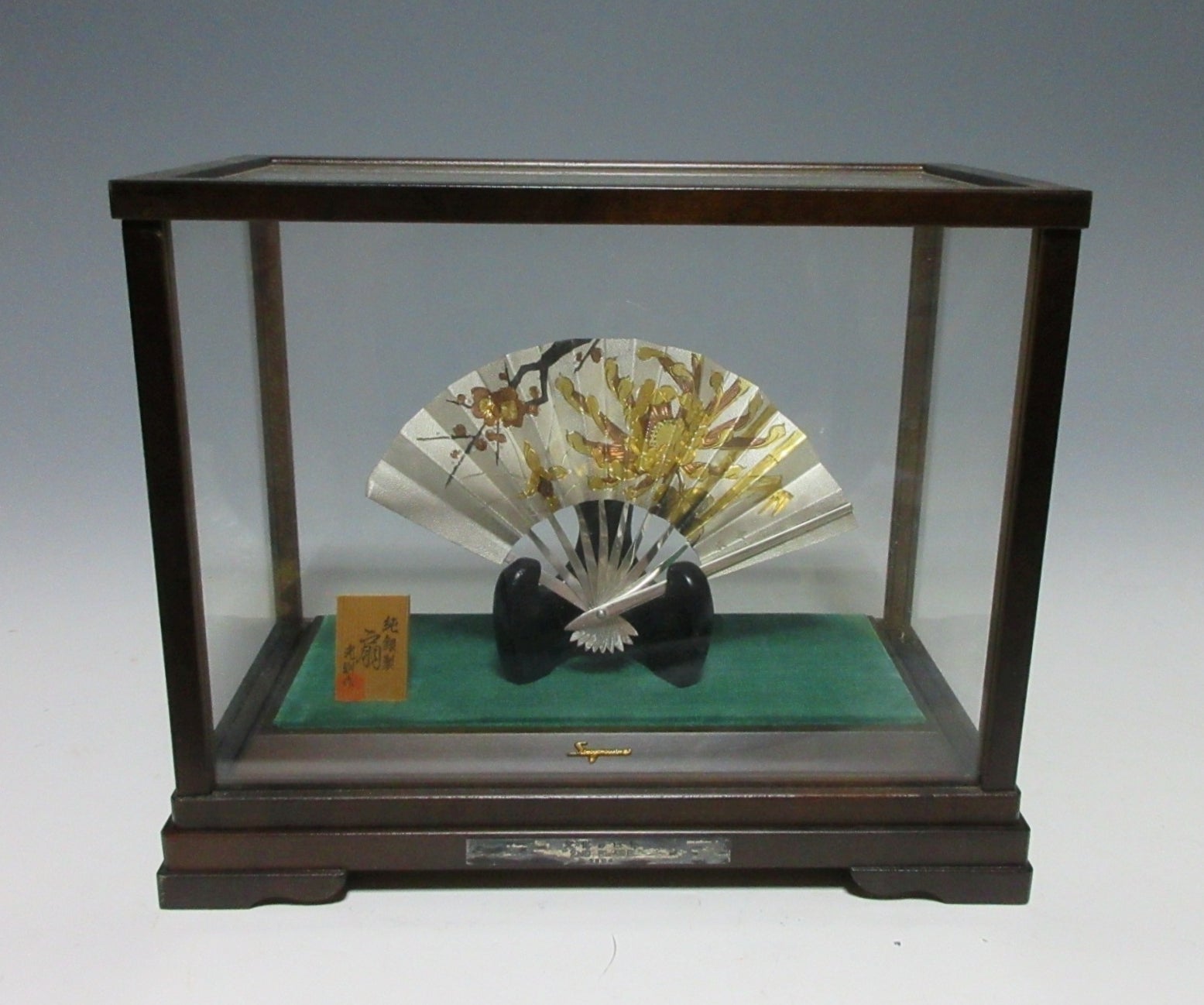 Jungin silver paper fan on a small stand inside a wooden-framed glass display case with a green base.