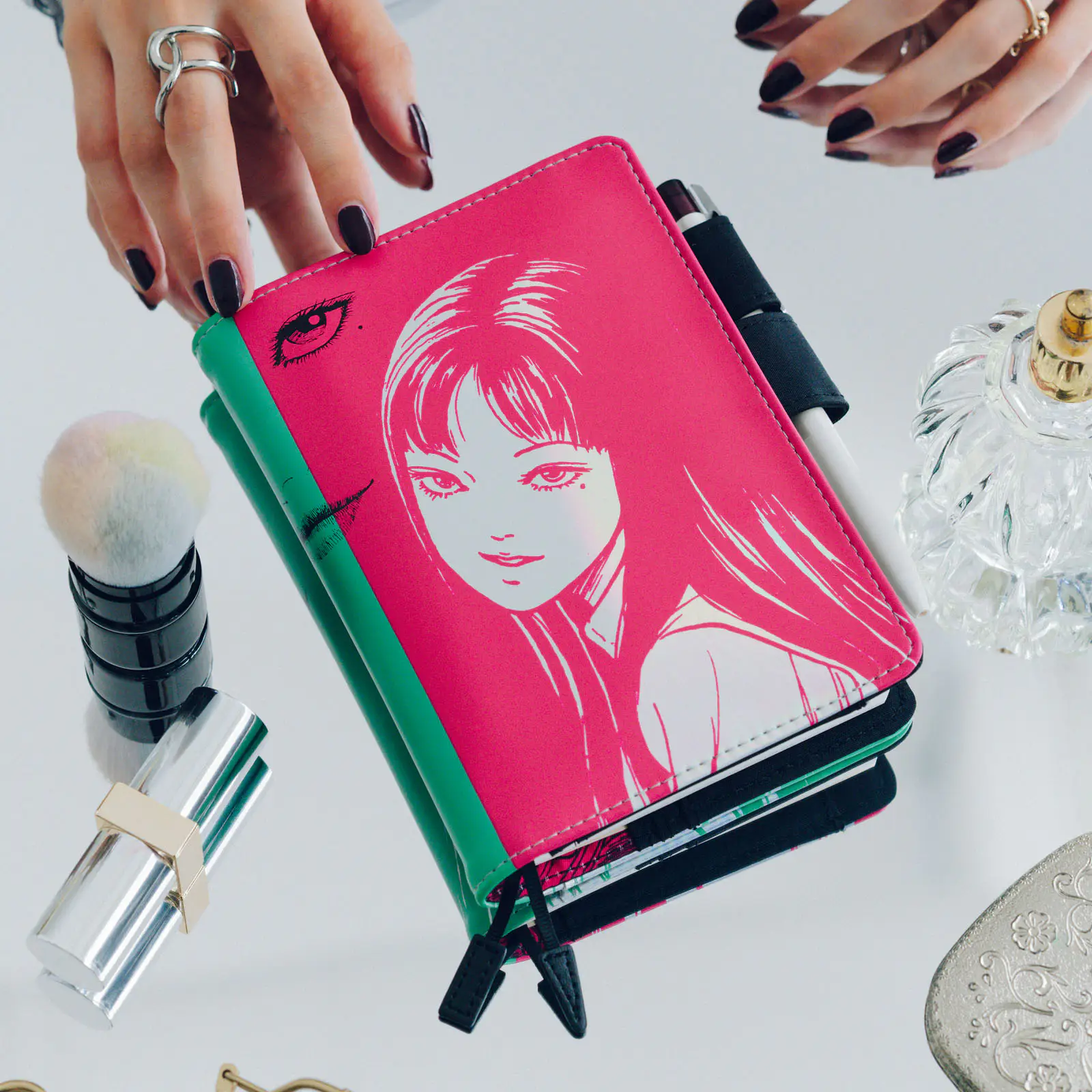 A pink and green A6 notebook with Junji Ito Tomie illustration on the cover held on a reflective surface beside makeup items.