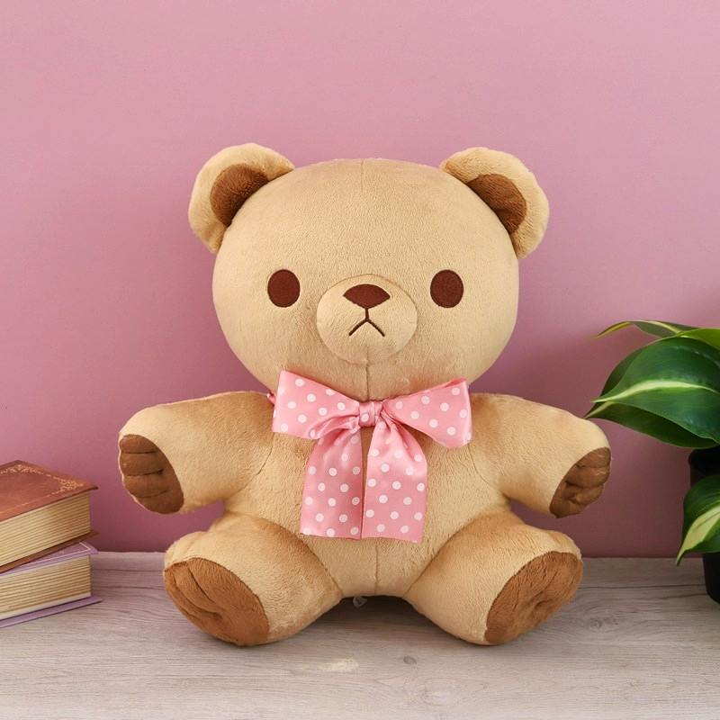 A Suzuki-san plush teddy bear with a pink polka-dot bow sitting against a pink background.