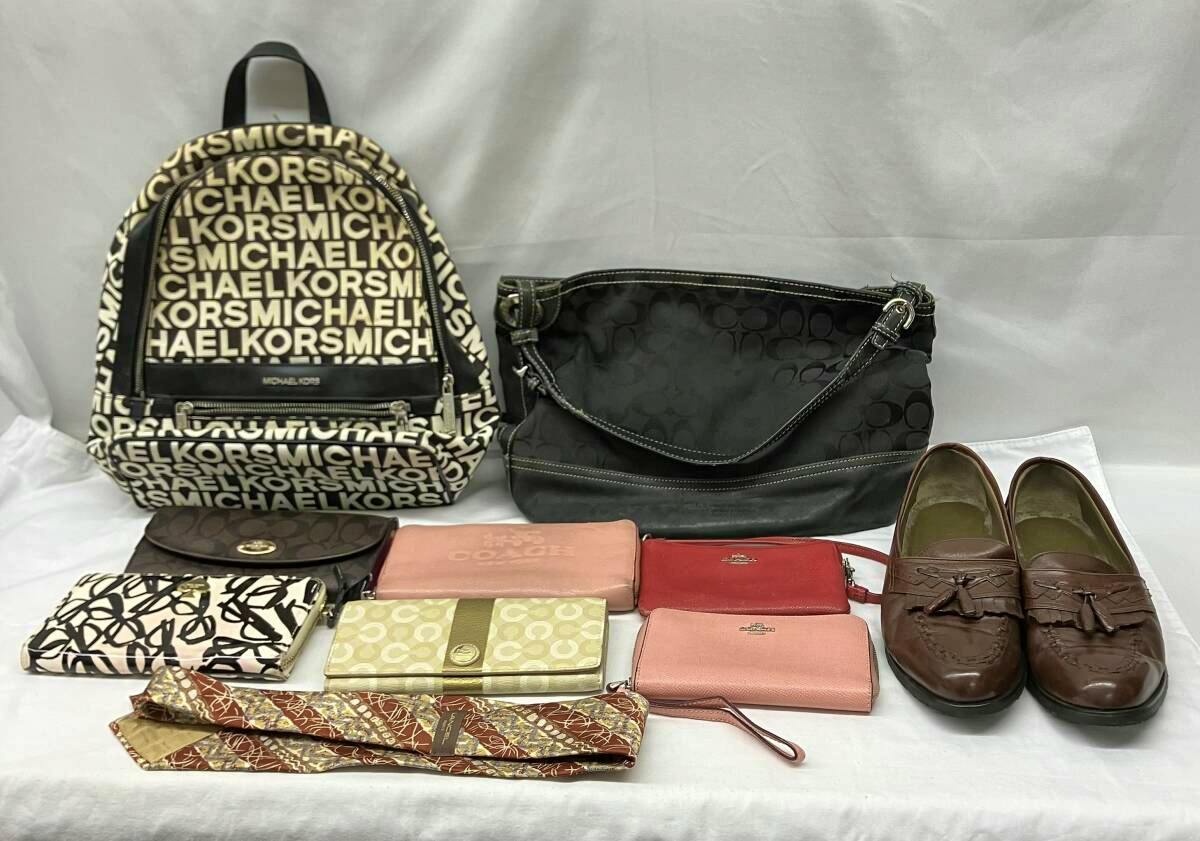 A group of branded bags, wallets, a pair of brown shoes, and ties arranged on a white background, with a Michael Kors logo-print backpack at left and a black Coach shoulder bag at center.