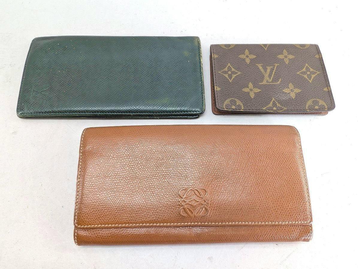 Three wallets on a white surface: a dark green wallet, a Louis Vuitton monogram wallet, and a brown Loewe long wallet.