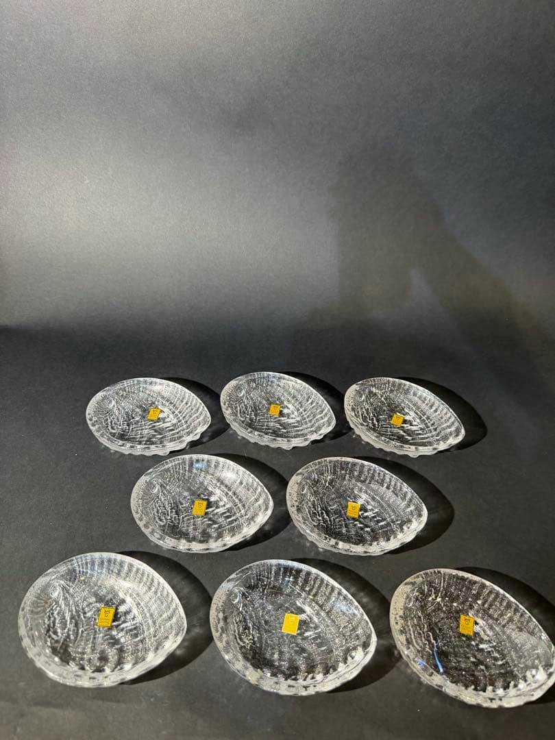 Eight shell-shaped clear glass small plates arranged on a dark background, each showing a small yellow sticker.