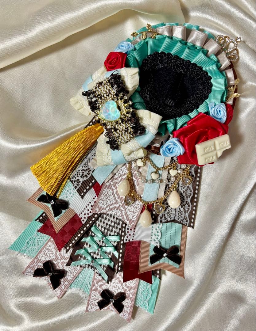 Front view of the embroidered brooch showing aqua-blue and brown rosette ribbons, red and light-blue rose accents, a gold tassel, a decorative bow, and layered patterned ribbon tails on a satin background.