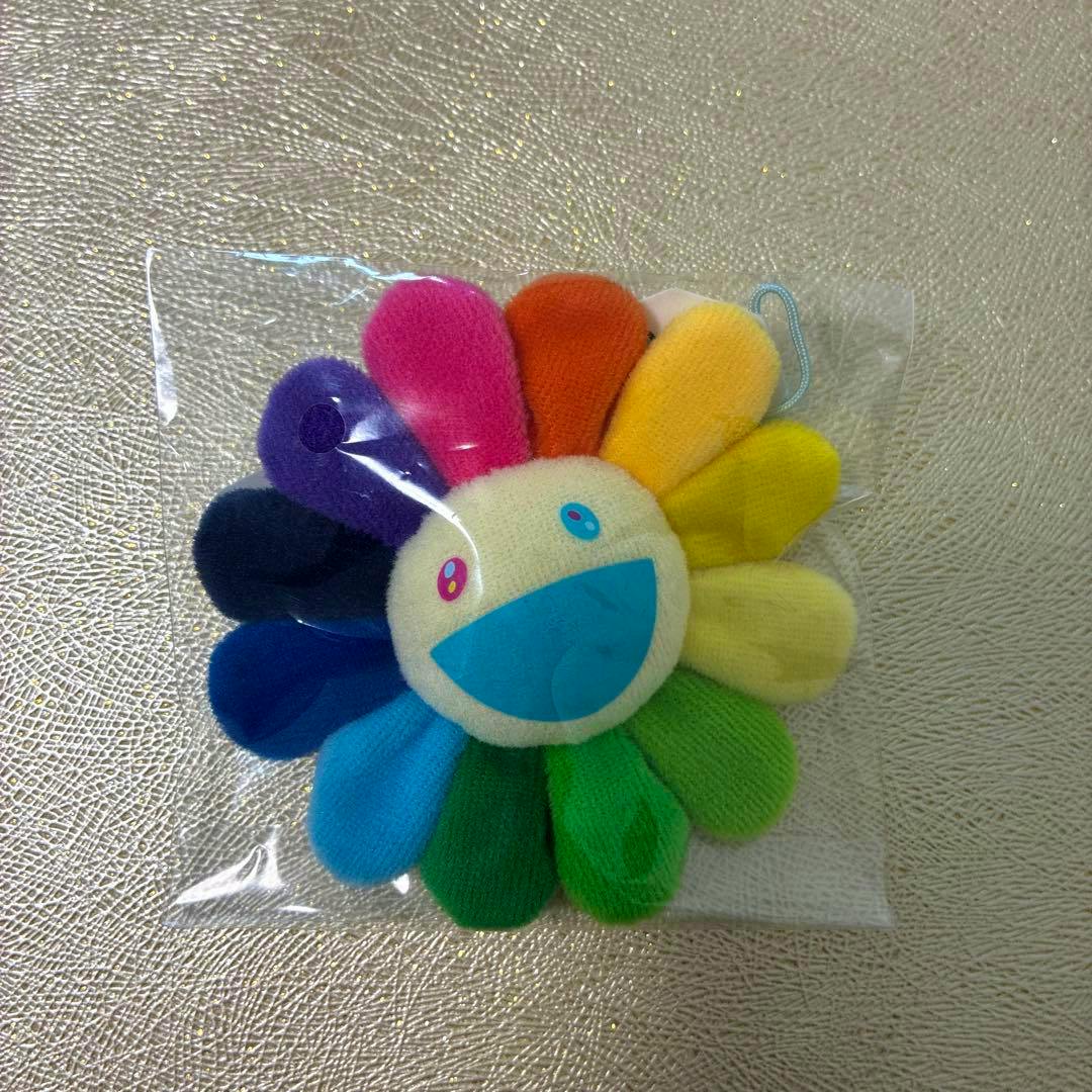Front of a multicolored Kaikai Kiki flower plush pin badge in clear plastic packaging on a textured background.