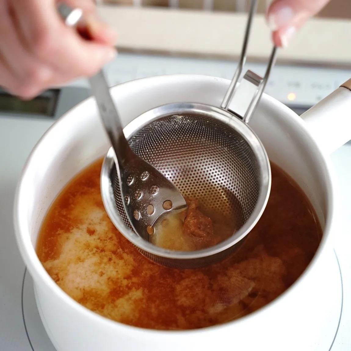 Stainless steel perforated miso strainer held over a pot of miso soup while a spoon stirs.