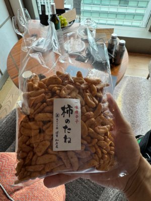 A hand holding a clear plastic bag of packaged 柿のたね rice crackers with a white label.