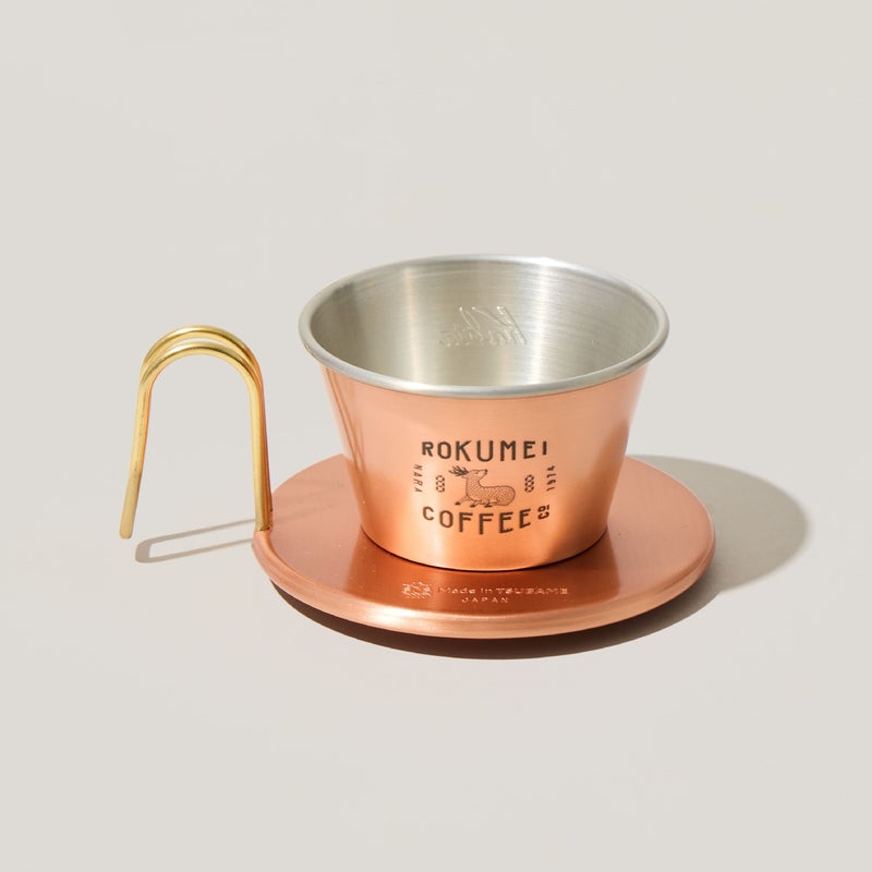 Copper Kalita wave coffee dripper with a gold handle and round base, showing printed branding, on a light background.