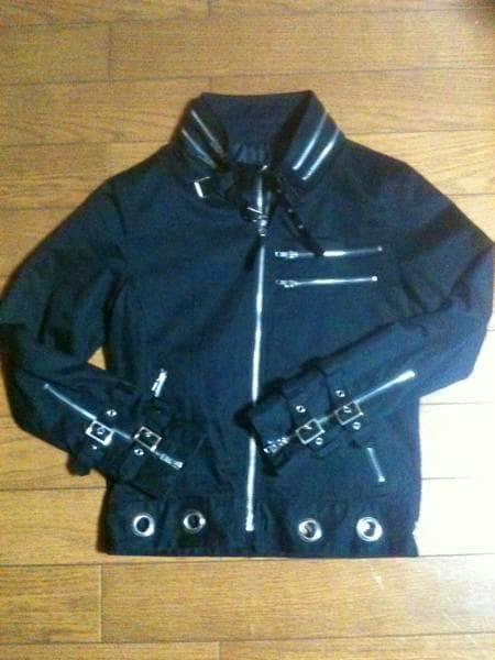 Black men's riders-style jacket (polyester) laid flat showing front zipper, chest zip pockets, buckled cuff straps, and metal grommets on the hem.
