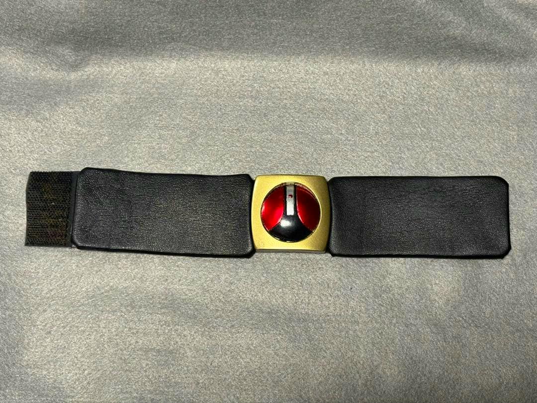 A Kamen Rider BLACK RX 1/1 wristbit prop replica with a gold square buckle showing a red-and-black circular emblem on a black strap.