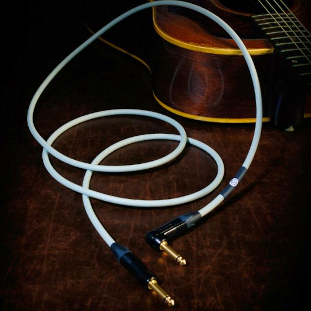A light-colored acoustic cable coiled on a dark surface with gold 1/4-inch connectors and part of an acoustic guitar visible in the background.